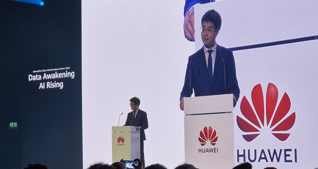 A man in a dark suit and glasses speaks at a podium with the Huawei logo. Behind him is a large screen showing his image and the event slogan: “Data Awakening, AI Rising.” An audience watches in a conference hall.